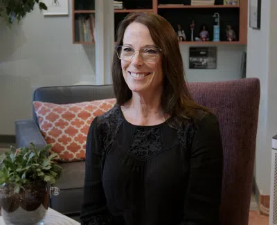 Smiling woman with glasses seated in a cozy, modern living room with plants and patterned pillow.