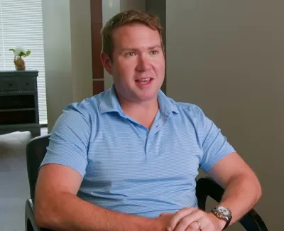 Man in blue polo shirt sitting in a chair inside a modern room with light walls and a window.