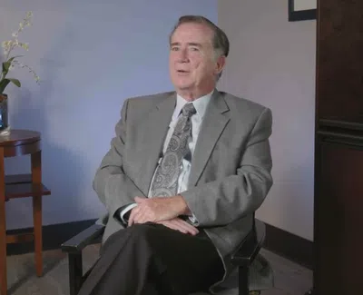 Man in gray suit sitting on chair in an office setting with a wooden table and white orchid plant.