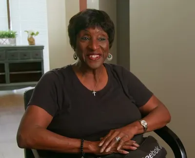 Smiling woman with cross necklace sitting indoors in casual black t-shirt, hands folded over a Reebok bag