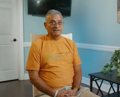 Man wearing orange shark t-shirt sitting on chair in blue room with plants and smile sign on table.