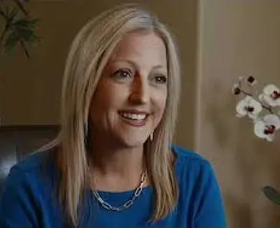 Smiling blonde woman in blue top sitting indoors with orchids and plants in the background