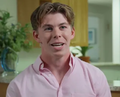 Smiling young man with light brown hair wearing a pink shirt indoors, with plants and kitchen background.