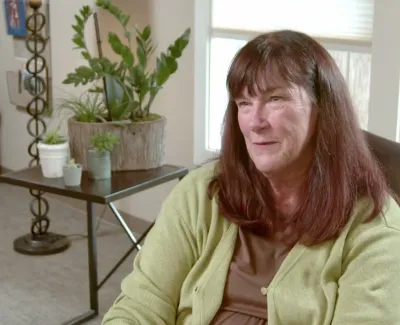 Middle-aged woman with brown hair in green cardigan smiling indoors with plants and chair in the background