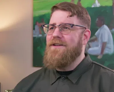 Bearded man with glasses smiling indoors near a lamp and colorful painting on the wall behind him