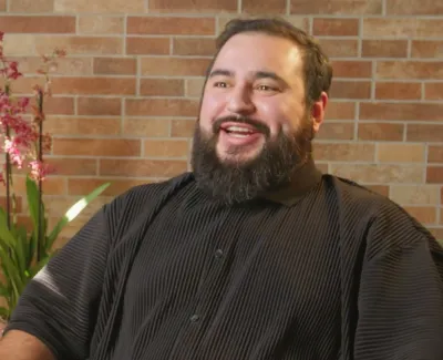 Smiling bearded man in black shirt sitting indoors with green plants and a brick wall background.