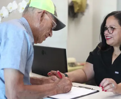 Senior man signing paperwork assisted by a smiling female professional at office reception desk.
