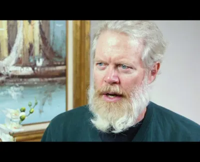 Middle-aged man with white beard and blue eyes speaking, with a painting and flowers in the background