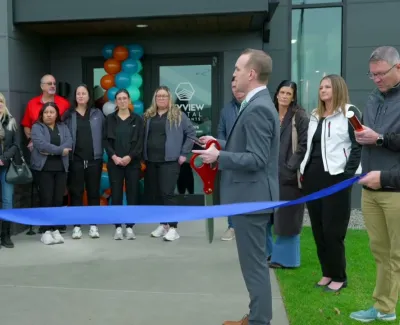 Group of people at a ribbon-cutting ceremony outside a dental office with balloons and large scissors.