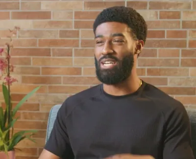 Man with beard and black shirt speaking in front of brick wall with indoor plants in background