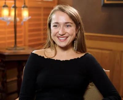 Smiling woman with long hair wearing black top sitting in cozy room with wooden walls and magazines on rack.