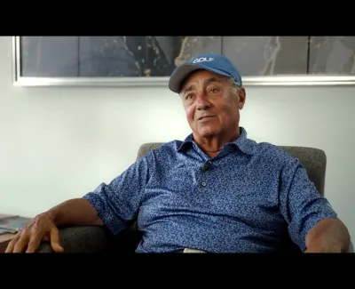 Elderly man wearing a blue golf cap and patterned shirt sitting and relaxing indoors on a chair.