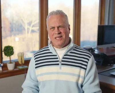 Man in a light blue striped sweater sitting in an office with large windows and wooden accents.