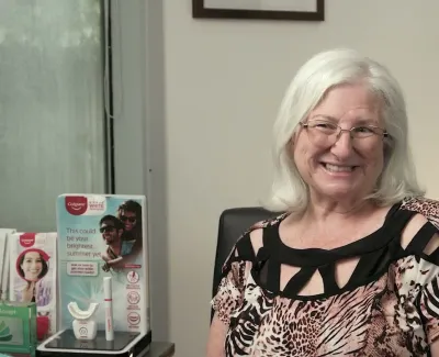 Smiling elderly woman with white hair and glasses sitting in dental office with promotional displays in background