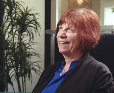 Smiling elderly woman with red hair wearing blue shirt and black sweater sitting indoors with plant background