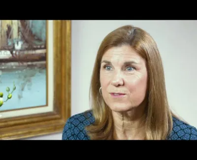 Middle-aged woman with light brown hair wearing blue patterned top sitting indoors near a framed painting and white flowers.