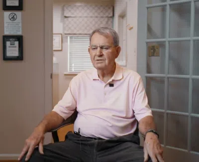 Elderly man in pink polo sitting in a chair indoors with neutral background and window blinds.