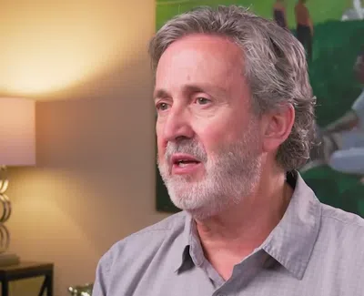 Middle-aged man with gray hair and beard speaking indoors with lamp and artwork in the background