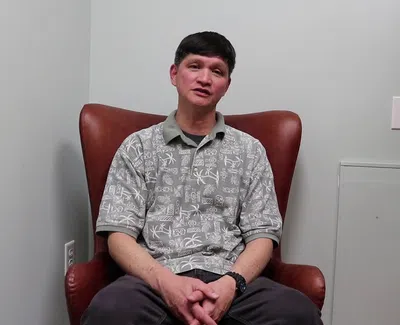 Man wearing patterned shirt sitting on brown leather chair against plain wall with plant on the side