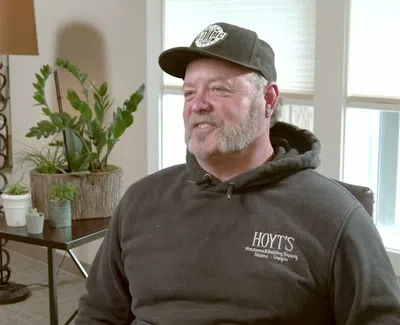 Man wearing a black Hoyt's hoodie and cap sitting in a room with plants and sunlight through windows