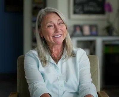 Smiling middle-aged woman with gray hair sitting in a cozy room with shelves and chairs in the background
