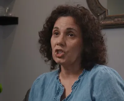 Woman with curly hair wearing a blue blouse speaking indoors next to a green plant and wall decorations.