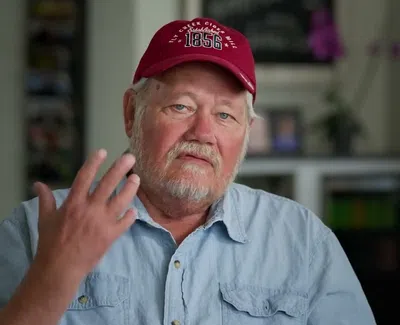 Older man with white beard wearing red cap and blue shirt gesturing with hand indoors.