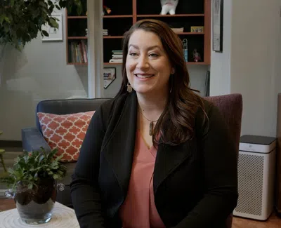 Smiling woman with long brown hair sitting in a cozy living room with plants and decorative pillows.
