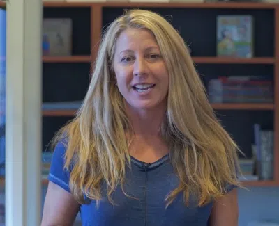 Smiling woman with long blonde hair in a blue shirt standing indoors with bookshelves in the background