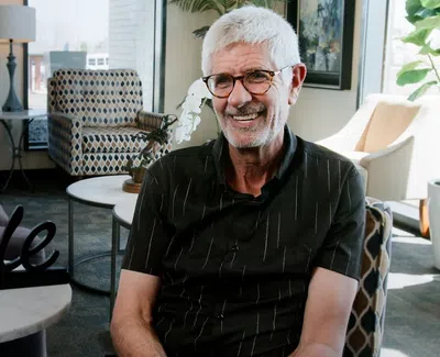 Smiling elderly man with glasses sitting in a modern living room with decor and a 'Smile' sign on a table.