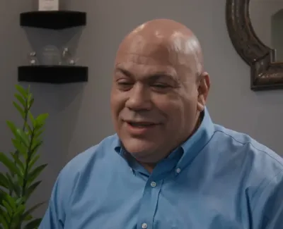 Smiling man in blue shirt sitting indoors with plant and decorative mirror in background