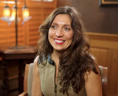 Smiling woman with long hair sitting indoors near wooden furniture and warm lighting.