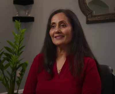 Smiling woman with long dark hair wearing a red top sitting indoors with plants and a mirror in the background