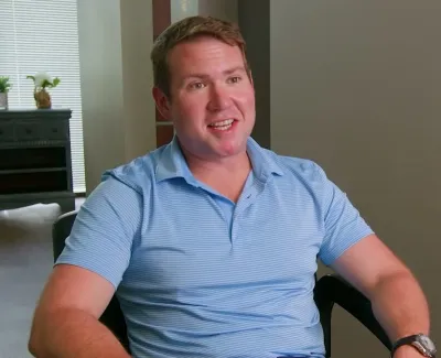Man in blue polo shirt sitting and smiling indoors near window with blinds and furniture in background