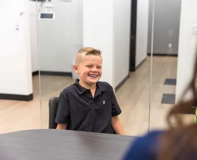 Smiling boy with braces sitting at a table in a modern office setting speaking with a person.