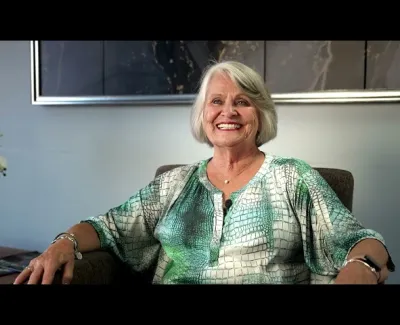 Smiling elderly woman with short gray hair wearing a green patterned blouse sitting comfortably in a chair.