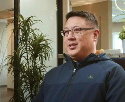 Man wearing glasses and navy zip-up jacket sitting indoors with plants and modern office background.