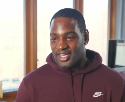 Smiling man in a maroon Nike hoodie standing indoors near windows with a small potted plant and computer in the background