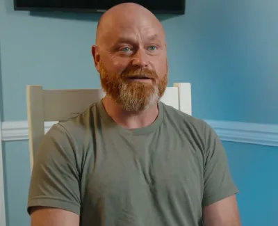 Bearded man with shaved head sitting in a chair against a blue wall with a plant nearby.