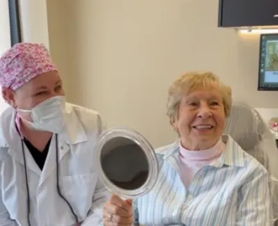 Smiling elderly woman holding a hand mirror with a healthcare worker in a medical setting wearing a mask and cap.
