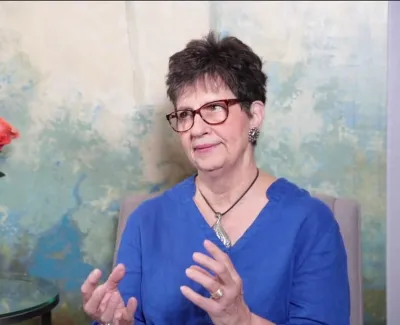 Woman in blue shirt gesturing while seated with vase of orange roses on glass table beside her