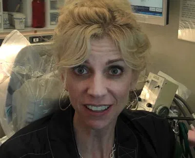 Smiling woman with blonde hair sitting in a medical or dental office surrounded by equipment and supplies.