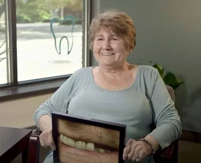 Elderly woman smiling and holding a photo of her teeth in a dental office with plants and tooth logo.