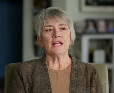 Middle-aged woman with short gray hair speaking indoors, wearing a brown top and lace cardigan in a cozy room.