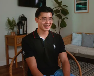 Smiling man wearing glasses and black polo sitting in a cozy living room with plants and framed art.