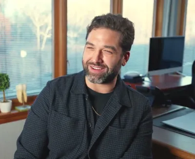 Man with beard and dark shirt smiling in a bright office with large windows and desk with computer.
