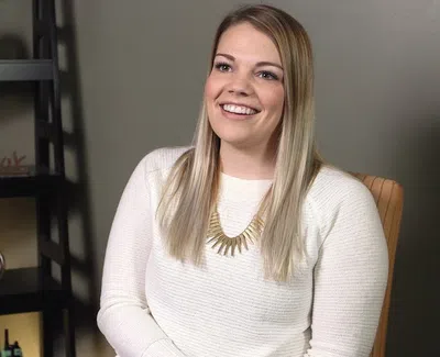 Smiling woman with long blonde hair sitting in a chair wearing a white sweater and gold necklace.