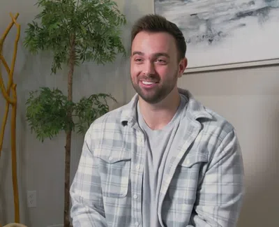 Smiling man in gray plaid shirt sitting indoors with decorative plants and wall art in background