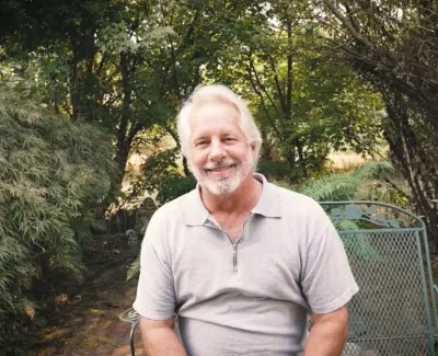 Smiling elderly man with white hair sitting on a metal chair in a lush green garden outdoors.