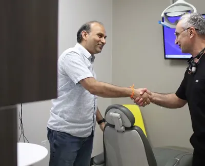 Dentist and patient shake hands in a dental office with computer screens showing dental images.
